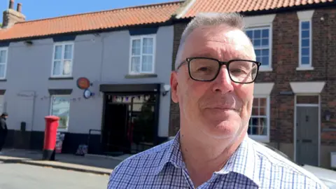A man with short light hair and black-framed glasses wears a blue-cheque shirt as he stands in front of a village shop and adjoining house.