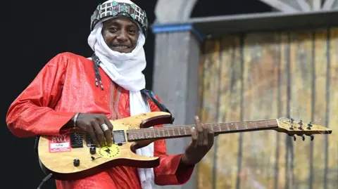 A man dressed in red smiles as he plays the guitar, looking straight into the camera.