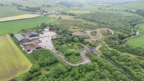 the site of an illegal dump sits surrounded by green fields and countryside. a number of dilapidated buildings are on the left, while minor roads run across the width of the site