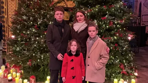 Heather Johnstone The four members of the Johnstone family standing in front of multiple Christmas trees. Heather and her two sons are dressed in coats and scarves, whereas the daughter is wearing the coat.