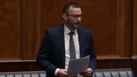 TUV MLA Timothy Gaston at the Northern Ireland Assembly. He is wearing a dark suit, white shirt and blue tie, as well as glasses. He is holding several sheets of A4 paper and is standing in the assembly chamber, behind him is a mahogany wall and a blue leather bench seat.