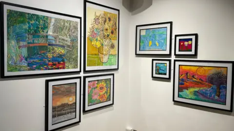 Amelia Riley/BBC News Eight pieces of artwork are displayed in the corner of a room, each one in a black frame. They vary in size and display natural themes such as flowers, mountains, and a river, in a variation of bright colours.