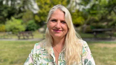 Victoria Oliver smiling at the camera. She is sitting in a park area with picnic benches, grass and trees in the background.
