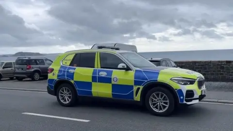 BBC A police car is driving along a coastal road with cars parked along the sea wall. The sea can be seen beyond the wall and part of the mainland in the distance.