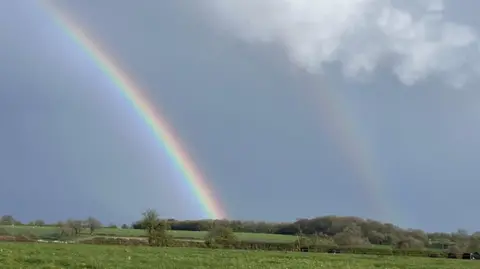 Cotswold Bob A double rainbow appears in a grey sky above fields of grass bordered by trees. One rainbow is much more distinct than the other