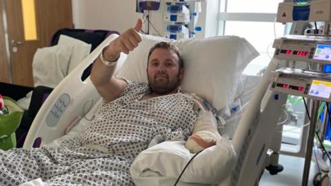 A picture of Jason taking in hospital. He is wearing a hospital gown and is smiling and giving a thumbs-up to the camera. His is attached to several different machines and his amputated left arm is shown heavily bandaged. 