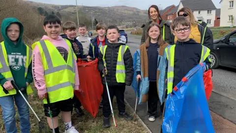 Eight schoolchildren pictured holding litter pickers and rubbish bags smile towards the camera. They are stood grouped together on the side of a road, many of them wear yellow high-vis vests. Two teachers can be seen stood smiling behind them. A residential estate can be seen in the background in front of some tree covered mountains. 