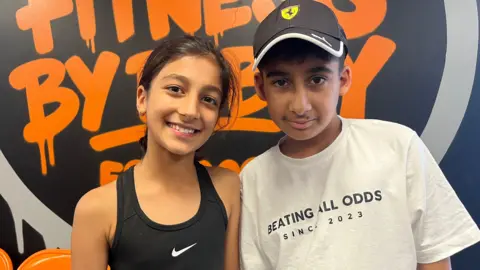 Isla is wearing a black Nike sports vest and is next to her borther Ariv who is wearing a white t-shirt and black cap. They are behind an orange and black Fitness by Bobby logo.