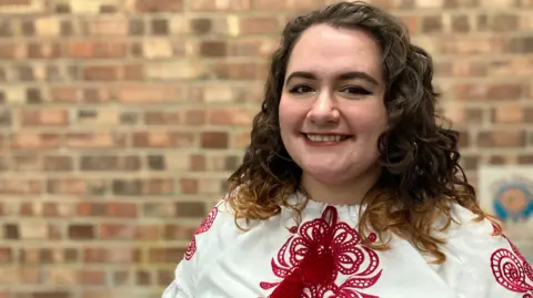 Yana Kozah is standing in front of a red brick wall. She is wearing a white shirt with red embroidery. Her hair is curly and brown. She is smiling at the camera.
