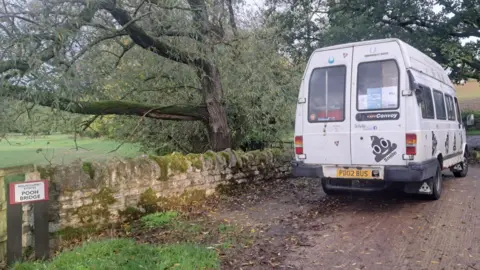 Barry Busby An old white minibus covered in poo emoji stickers is on a bridge. A sign to one side reads "Pooh Bridge".