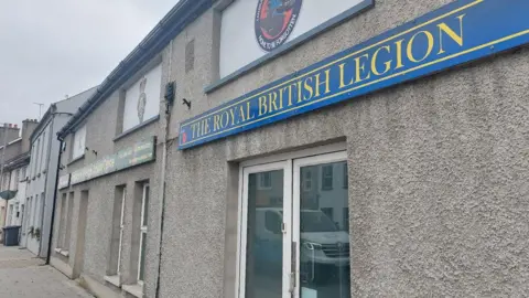 A picture showing the Royal British Legion building in Tandragee. It is a grey building with white doors, with a sign above it. The sign is blue with white writing that says 'The Royal British Legion'.