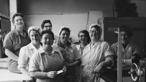 Black and white photograph of eight women posing to the camera in a factory room. They are all smiling. Some are wearing head coverings. Others are wearing aprons. Most are wearing jumpsuits. 