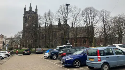 Photograph of a town centre car park in Stockport. The image shows several cars and in the background is St Mary's Church.