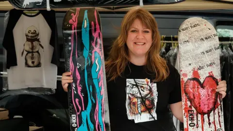 Craig Spencer Photos A woman with red hair stands in a stall where she is selling items. She holds two skateboards which have been painted on.