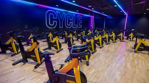 Everyone Active A room inside the centre with rows of many  yellow and black cycle machines. The ceiling has purple lighting and the floor is wood or wood laminate. The word CYCLE is emblazoned on the wall.