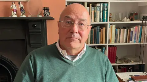 Mr Gunnell looks at the camera with a serious expression. He is bald and clean shaven, and wears frameless square glassed, a white shirt and a green v-neck jumper. He is in a study, and there are lots of books and a fireplace behind him. 