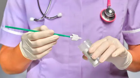 A nurse in a purple uniform places a sample, on the end of a green stick, into a clear and colourless tube. She has a stethoscope around her neck and wear white gloves.  