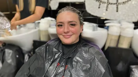 Abi Smitton / BBC News A woman at a hairdressers, with wet hair covered in conditioner. She's sitting in front of a sink and smiling at the camera. She's wearing a black cape and a bin bag over the top
