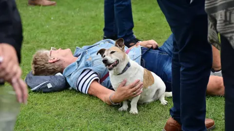 Pacemaker Press A dog is sitting next to a woman who is lying on grass. She has her hand on the dog. People are standing around them. 