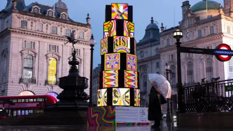 A brightly lit, multi-coloured cube sculpture stands in Piccadilly Circus at dusk, with a person holding an umbrella on wet pavement and historic buildings and a London Underground sign in the background.