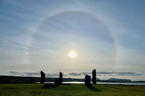 Holly Elson Standing stones in Orkney with halo of light overheard, and sea and land in the distance, under a cloudy blue sky.