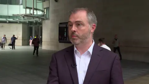 Neil Garratt in a purple suit speaking during an interview outside the BBC Broadcasting House.
