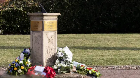 Crown Copyright 2025 A stone sundial has flowers, including a wreathe of poppies, laid next to it. Gold plaques on each side of the sundial lists names of people killed in the bus crash.