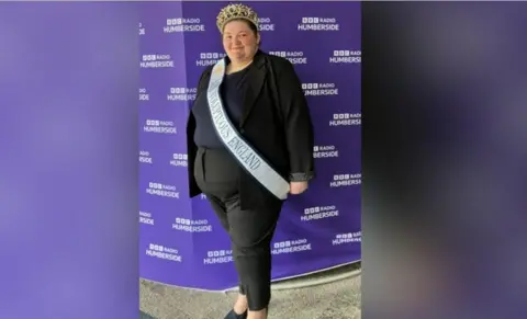 A full-length image of Philippa Mackinder, showing more of her sash and her outfit, which includes a black blazer over a navy top and black trousers and shoes. Her sash reads "Miss Voluptuous England". She is wearing a gold crown on her head. She is standing in front of a purple BBC Radio Humberside backdrop.