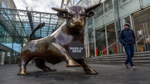 The Bullring bull is prominent on the left and in the middle of the photo. A man in blue on the right is looking at it.