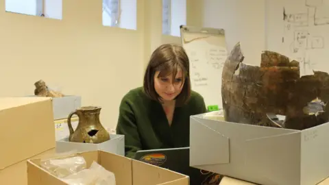Durham University/The Auckland Project Doctoral Researcher Emily Walker typing on a laptop as she carries out some post-excavation work. She is surrounded by cardboard boxes. Some of the boxes are open, revealing metal and gold looking artifacts inside them. One of the items resembles a vase. Ms Walker is wearing a green cardigan. Her straight brown hair reaches her shoulders and she has a fringe. There is writing on the whiteboards behind her. 