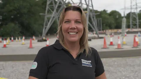 woman in the foreground wearing a black polo shirt with a “Morgan Sindall Infrastructure” logo, standing at an outdoor electrical infrastructure site and facing the camera. Behind the person are large steel electricity transmission structures, overhead power lines, and a gravel-covered work area. Numerous orange-and-white traffic cones mark pathways and restricted areas across the site. Trees line the background, and a blue site container is visible on the right. No bags or bins are visible.