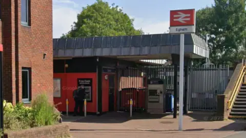 A Google Street image of the outside of Egham Railway Station. There is a sign with the railway logo and the word "Egham" under it.