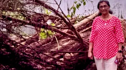 Irie Perera is wearing a red and white top, with white trousers. She has glasses on and is stood next to fallen trees.