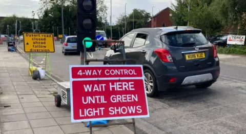 Sharon Edwards/BBC A red traffic sign says "4-way control - wait here until green light shows". The light is on green and cars are weaving through past another yellow sign that says "Dysart road closed ahead follow diversion".