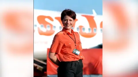 ITV Jane smiles at the camera in front of a plane. She wears an orange short sleeved shift and black trousers, and has short brown/red hair.