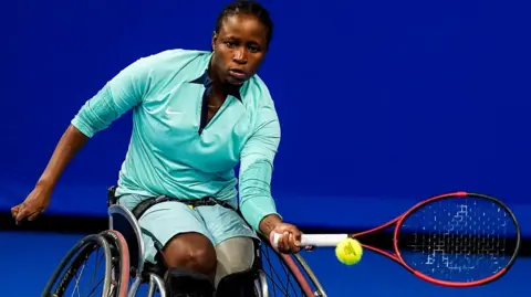 Kgothatso Montjane, wearing a pale green top and light blue shorts, stretches a tennis racket held in her left hand towards a tennis ball while she plays wheelchair tennis