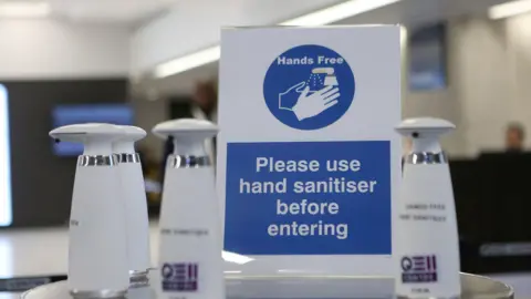 PA Media A table with hand sanitiser products sitting on it. A sign reads Hands Free, with a picture of the sanitiser spraying onto hands without being touched, and text reading PLEASE USE HAND SANITISER BEFORE ENTERING.