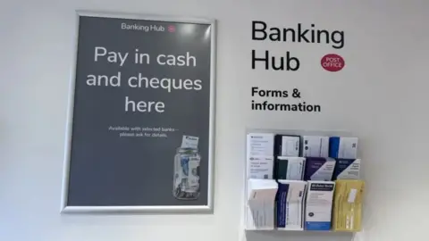 A grey sign with white writing saying "pay in cash and cheques here" on a white wall next to a black sign saying Banking hub above three rows of banking envelopes