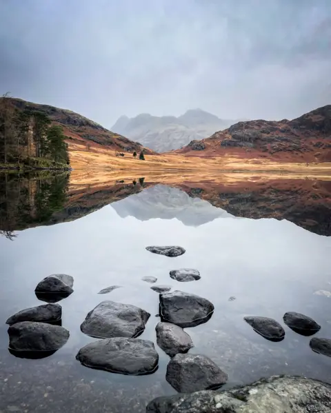 Adrian Conchie A perfectly still tarn, with a yellow and green hill beyond and distant craggy mountain reflected perfectly.