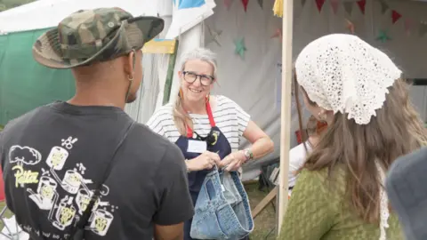BBC Clare seeking repairing a denim skirt in front of two festival attconcludeees. She is wearing an apron and striped t-shirt.