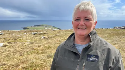 Head and shoulders shot of Elizabeth Johnson, who has short grey hair, smiling at the camera. She is wearing a grey t-shirt and a darker grey jacket with a navy "Saxavord UK Space Port" patch. She is standing on a shoreline and the sea is visible under grey skies in the background