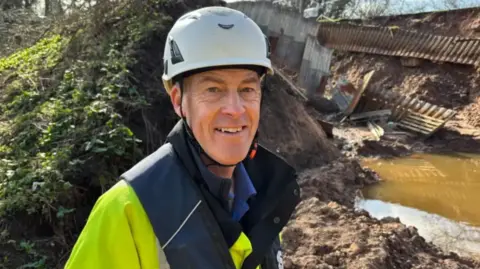 A man with a yellow and blue reflective jacket and a white helmet beside a brown embankment with brown water behind him
