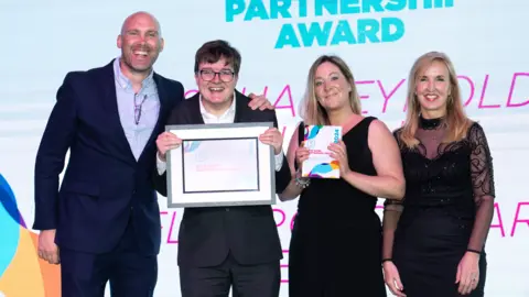 Weston College Josh Reynolds-Hailing wearing a black suit and white shirt, being presented with an award. BBC Radio presenter and actor Joe Sims is the left of picture, wearing a black suit and light blue shirt. Charlene Pickles and Jacqui Ford stand to the right of the picture, both in black dresses. All four in the picture are smiling at the camera.