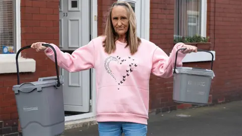 Tracy Harries stands in a street of red brick terraced houses with white upvc doors and windows. She is wearing a pink sweatshirt with a heart emblem and pale jeans and has long straight fair hair and is holding two grey food waste collection caddies out to each side