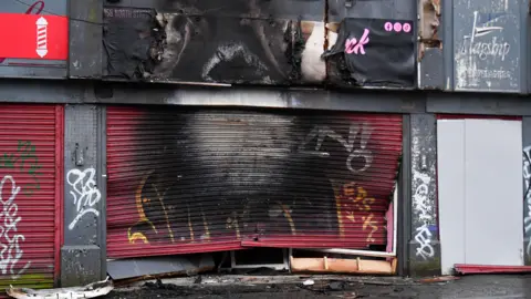 Pacemaker A shop front with red shutters is caved in with significant fire damage visible with black and white marks across it. 