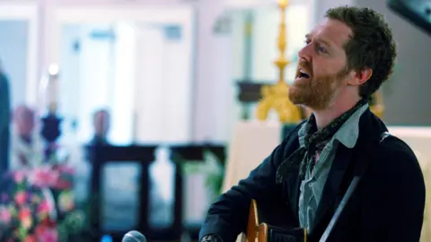 Glen Hansard has short curly reddish/brown hair and a ginger beard and is playing a guitar and singing in a church
