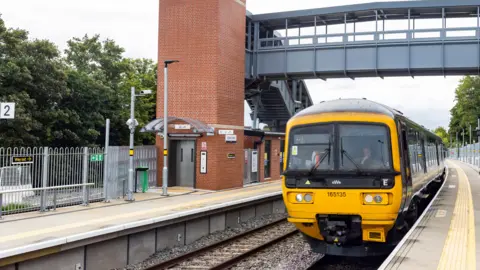 A train in the station at Ashley Down railway. The yellow front of the driver's compartment can be seen, with a driver inside. There is a grey footbridge over the train.