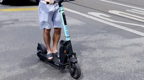 The bottom half of a male rider on an adult electric scooter.  He is wearing shorts and flip flops.