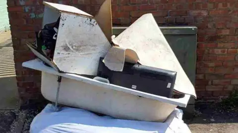 Hull City Council A thick white sheet with what appears to be a bathtub dumped on top of it in front of a brick wall. It is at an angle and has a TV in it as well as several pieces of wood