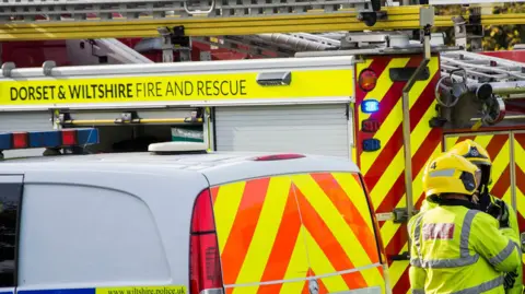 A police van parked in front of a Dorset and Wiltshire branded fire engine, both covered in prominent high-vis markings. Two firefighters can be seen speaking to each other, though their faces are not visible. 